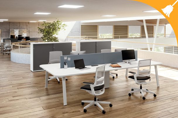 Modern open-plan office with wooden floors, white desks, office chairs, laptops, and dividers; potted plant and natural light in the background. A broom icon is in the top right corner.