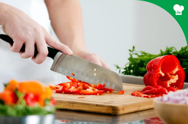 A person slices a red bell pepper with a chef’s knife on a wooden cutting board, surrounded by vegetables and greens.