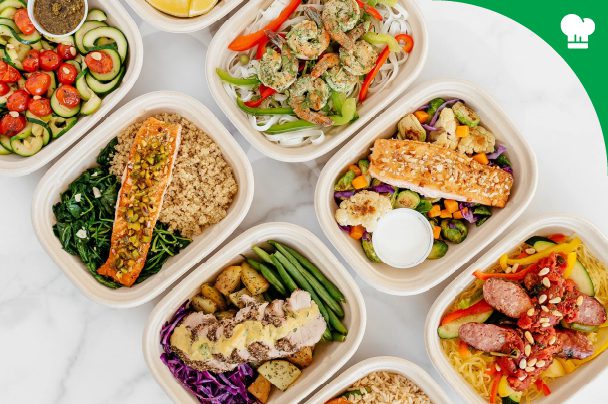 An assortment of prepared healthy meals in containers, featuring salmon, grilled chicken, shrimp, sausage, vegetables, and grains, arranged on a white surface.