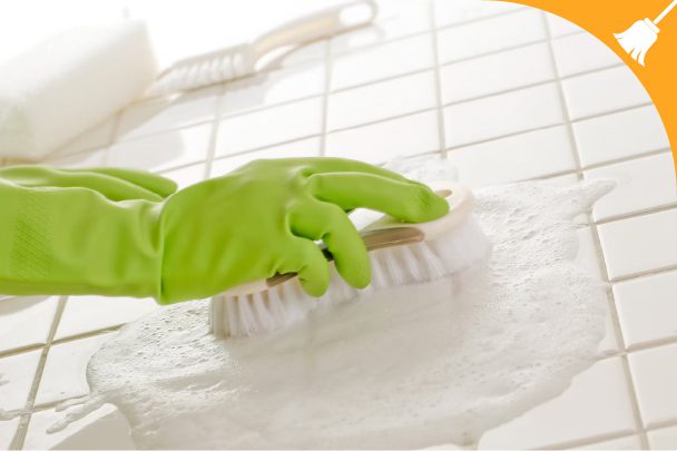 A person wearing green gloves scrubs soapy tiles with a brush, cleaning a tiled surface. A broom icon appears in the top right corner.