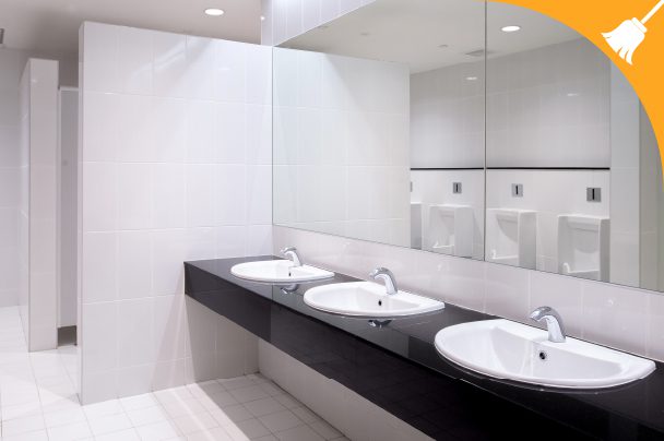 A clean public restroom with three white sinks, a large mirror, white tiled walls and floors, and urinals visible in the background. An orange corner features a white broom icon.