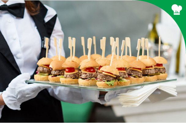 A server in formal attire holds a tray of mini burgers with skewers, next to a stack of napkins.