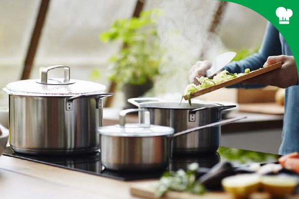 Person adding chopped vegetables into a pot on a stovetop, with two other pots nearby, in a bright kitchen setting.