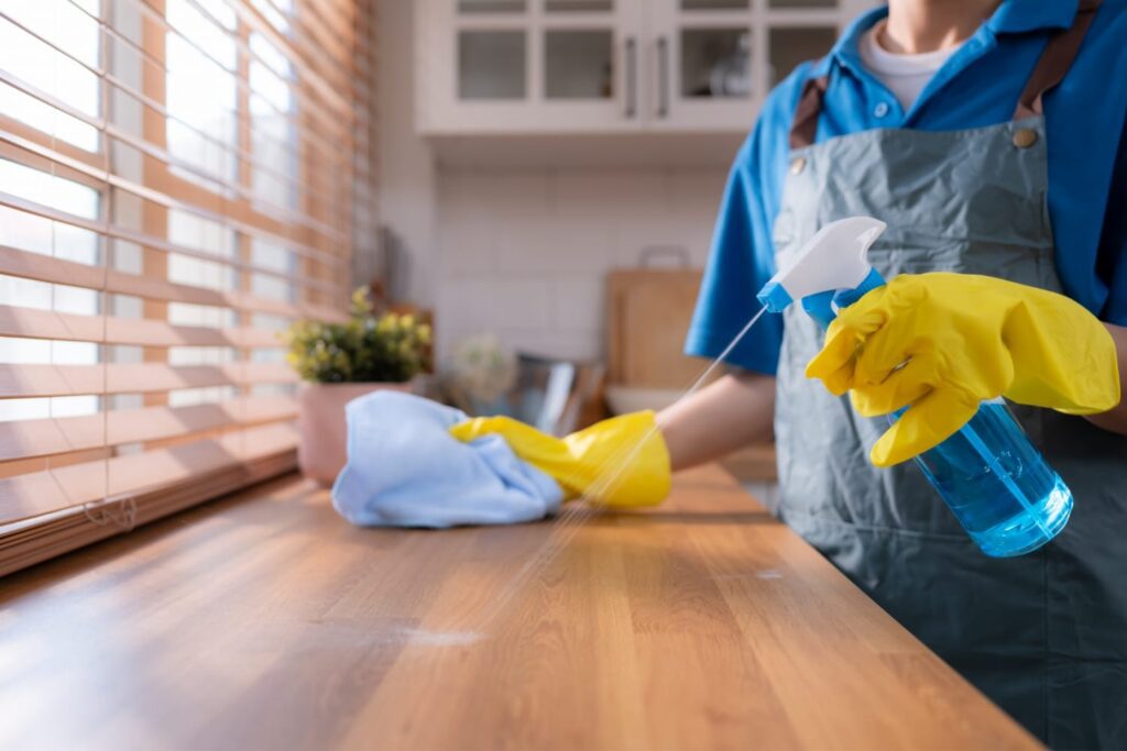 Person wearing yellow gloves cleans a wooden countertop with a blue spray bottle and a cloth in a bright kitchen.