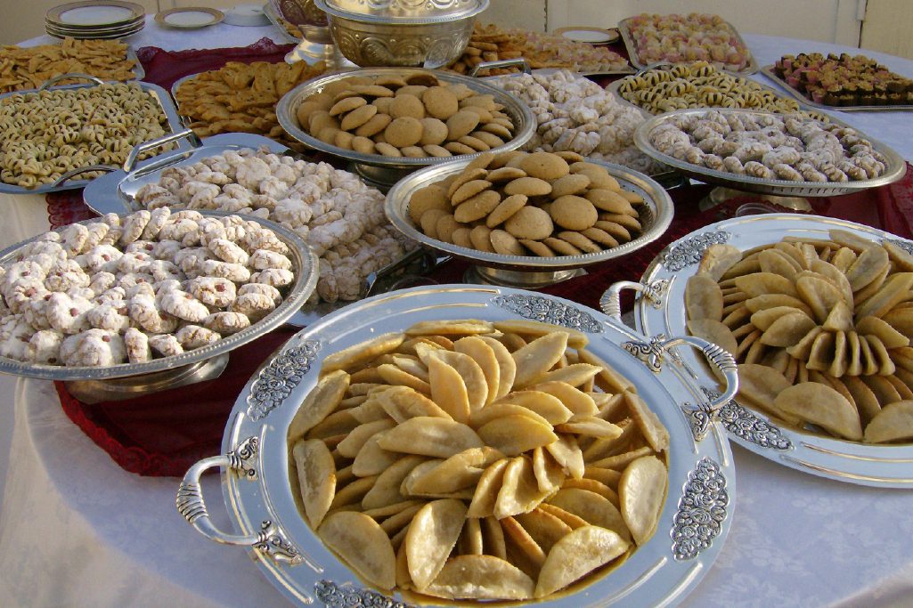 A variety of traditional Middle Eastern cookies and pastries are displayed on silver trays arranged on a table.