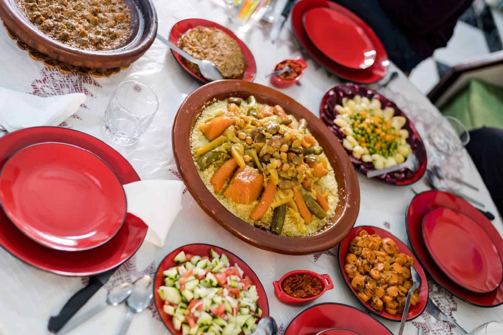 A table set with a large dish of couscous and vegetables, surrounded by various salads, stews, and red plates on a white tablecloth.