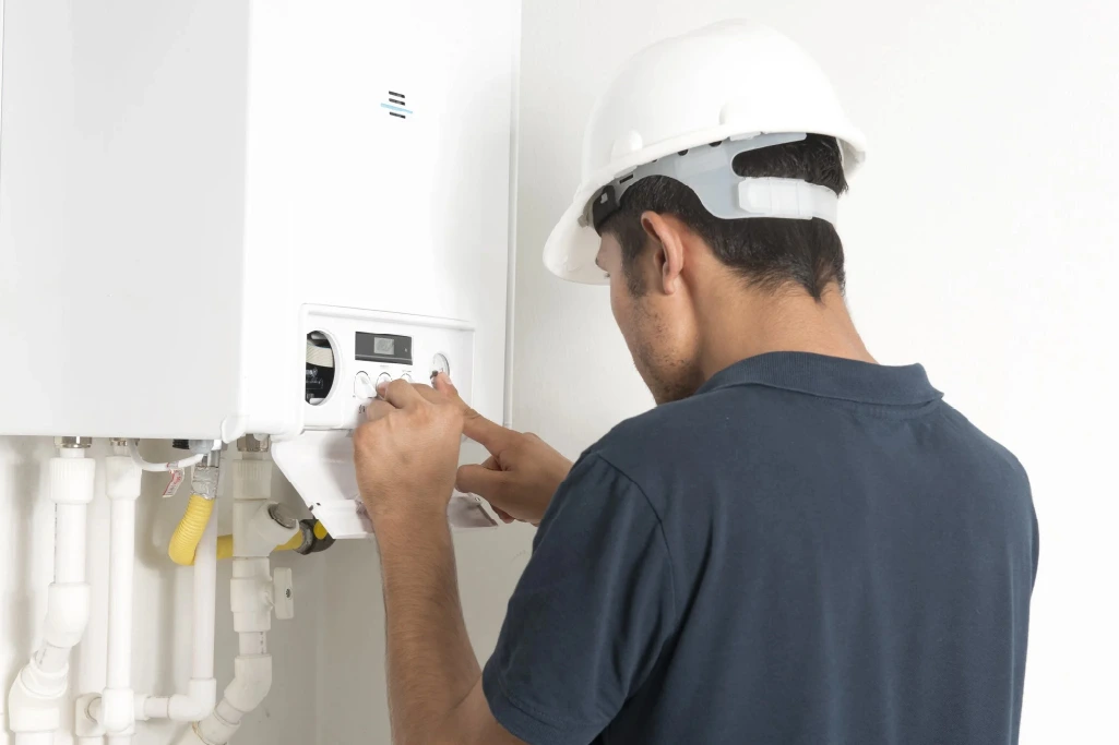 A worker in a hard hat adjusts the settings on a wall-mounted boiler or water heater.