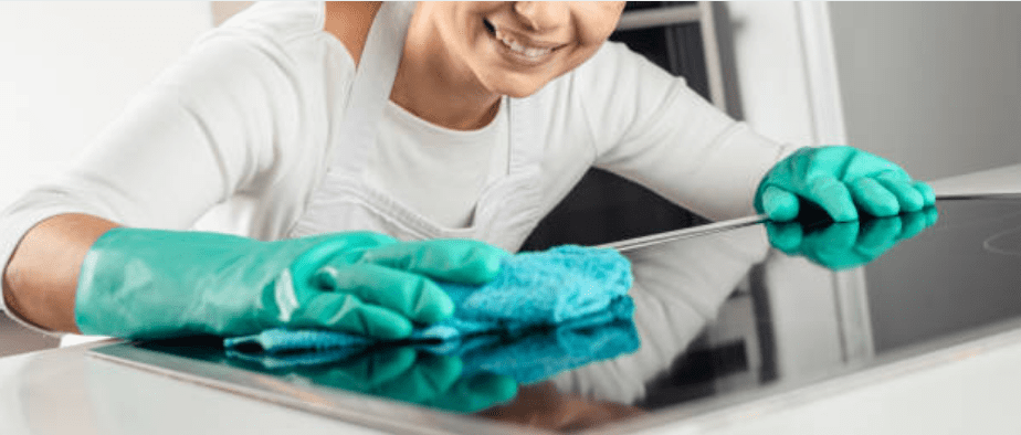 Person wearing green gloves and a white apron cleaning a kitchen stovetop with a blue cloth.