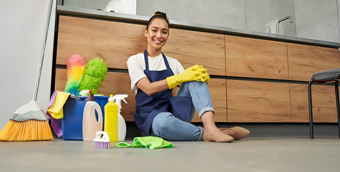 A woman wearing gloves and an apron sits on the kitchen floor, surrounded by various cleaning supplies and tools.