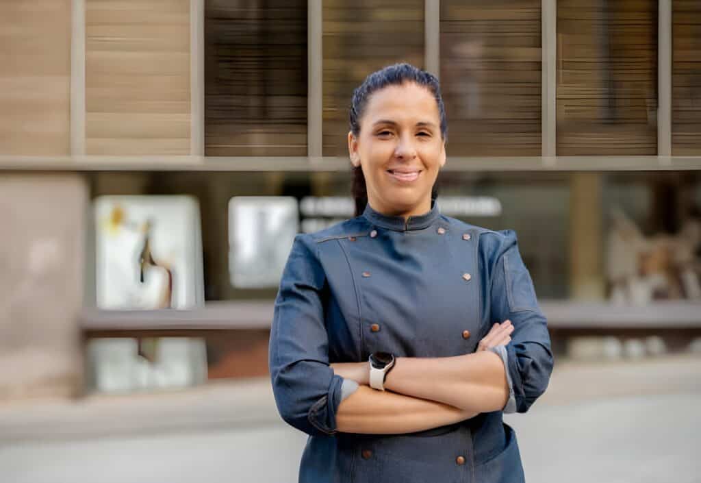 A cuisiniere in a chef’s jacket stands outside with her arms crossed, smiling, in front of a modern building.