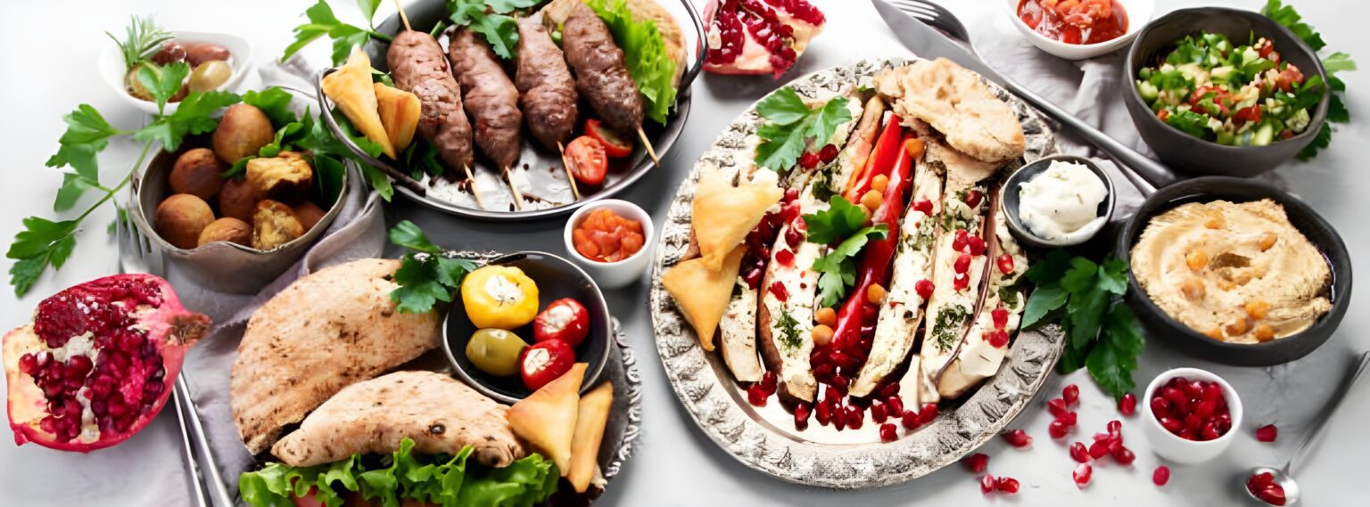 A cuisiniere’s spread of Middle Eastern dishes including pita bread, hummus, grilled kebabs, stuffed eggplant, fresh salad, olives, samosas, and pomegranate arranged artfully on a white table.