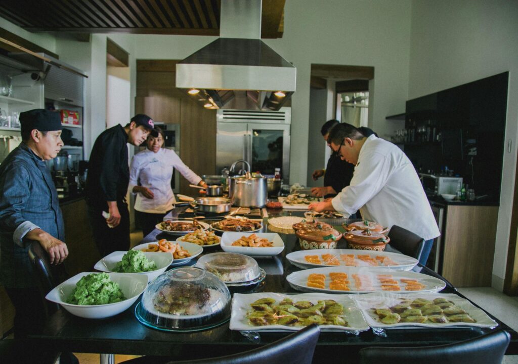 Several chefs and a cuisiniere prepare and arrange a variety of dishes on a large kitchen counter in a modern, well-lit kitchen.