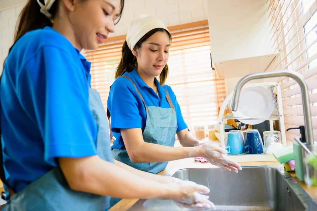 Two people in blue uniforms and aprons wash their hands with soap at a kitchen sink, with dishes and cups on a drying rack nearby.