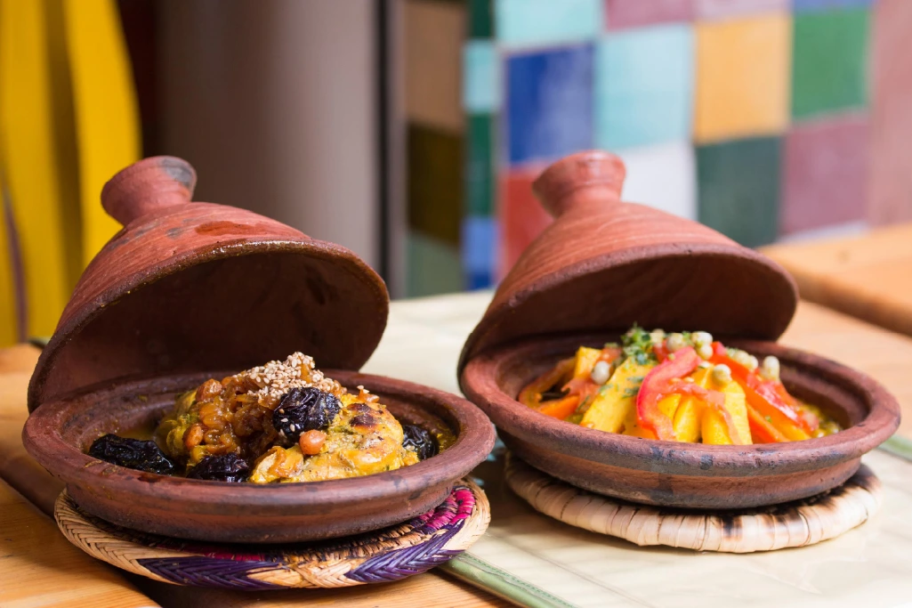 Two traditional Moroccan tagines with conical lids, one containing a meat and dried fruit dish, the other filled with vegetables, set on woven mats next to a colorful tiled wall.