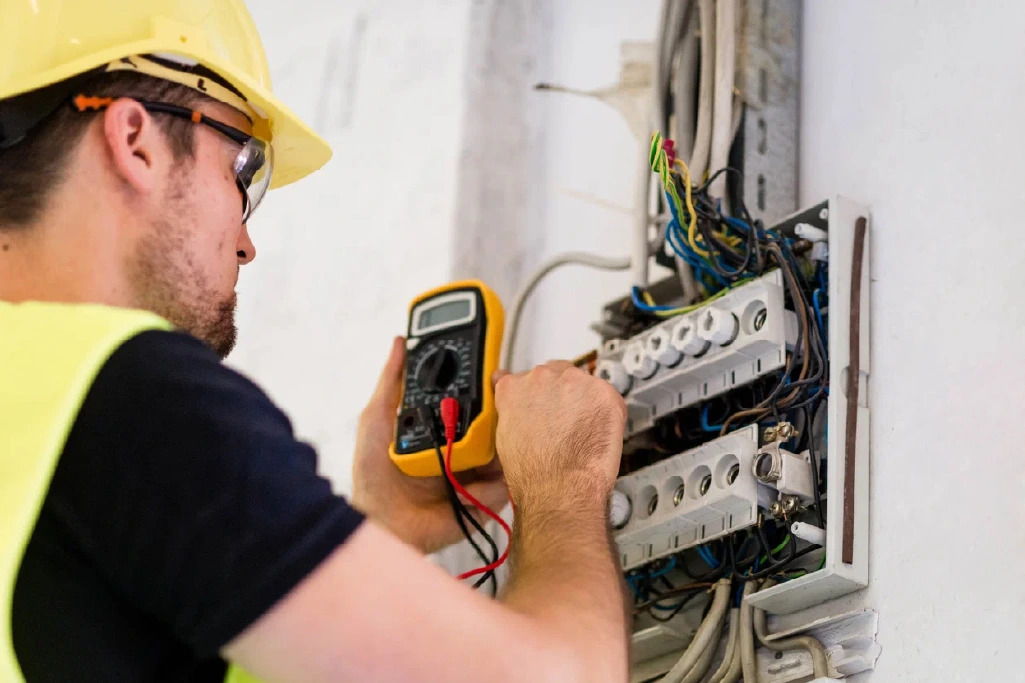 A person wearing safety gear uses a multimeter to test wires in an open electrical panel.