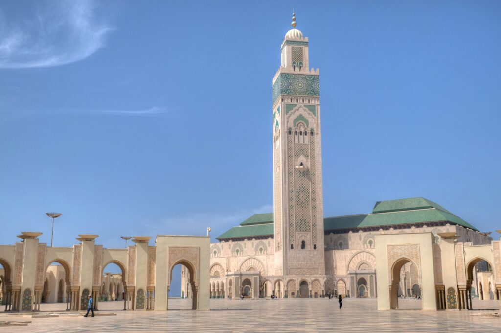 The image shows the Hassan II Mosque in Casablanca, Morocco, with its large courtyard and tall minaret. In the foreground, a cuisiniere prepares traditional Moroccan dishes as people walk around.