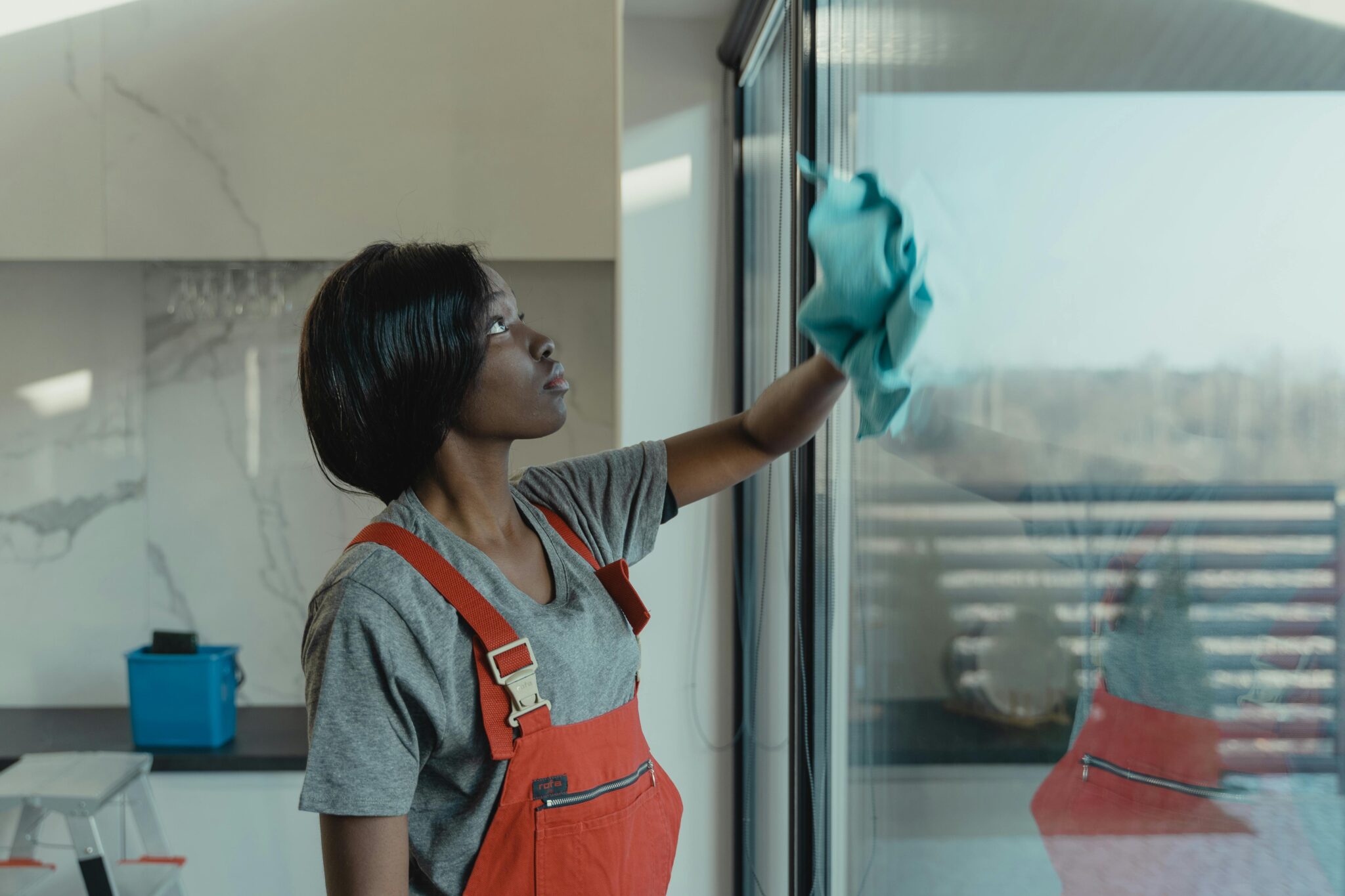 A woman wearing red overalls wipes a large glass window in a modern kitchen with a blue cloth.