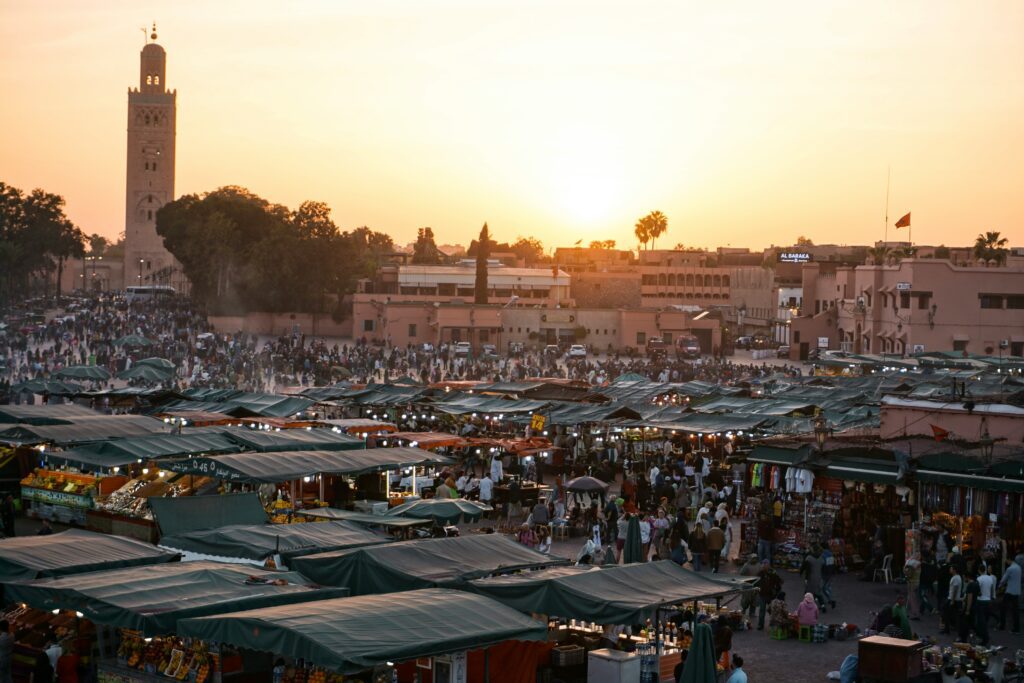 A busy outdoor market with many stalls and crowds at sunset, where a skilled cuisiniere prepares local dishes, set against the backdrop of a tall minaret and surrounding buildings.