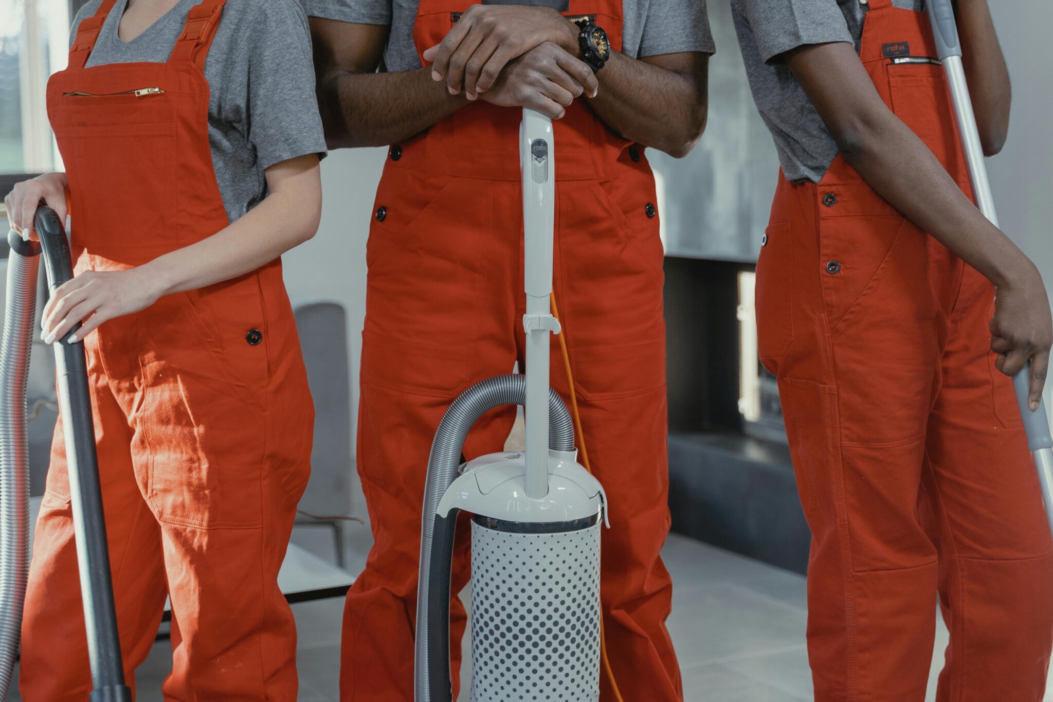 Three people in grey shirts and orange coveralls stand indoors holding cleaning equipment, including a vacuum cleaner and a mop.