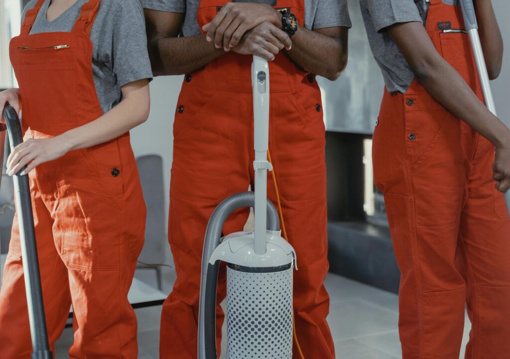 Three people wearing grey shirts and orange overalls stand indoors holding vacuum cleaners, shown from shoulders down.
