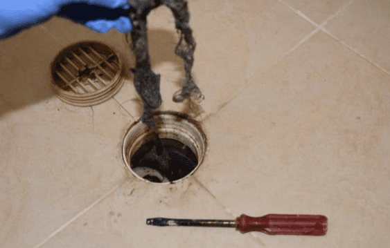 A gloved hand holds debris removed from a floor drain, with a drain cover and a screwdriver lying on the tiled floor nearby.