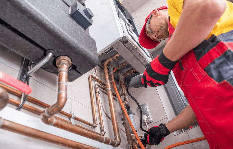 A plumber in a red uniform inspects and works on copper pipes connected to a heating unit on a tiled wall.