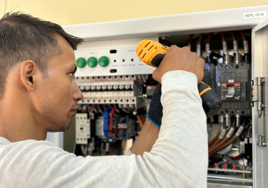 An electricien à Marrakech uses a cordless drill to work on an open electrical control panel filled with various wires and switches.