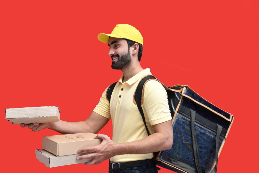 A delivery person in a yellow cap and shirt, reminiscent of a Société d'électricité uniform, carries pizza boxes and a delivery bag against a plain red background.