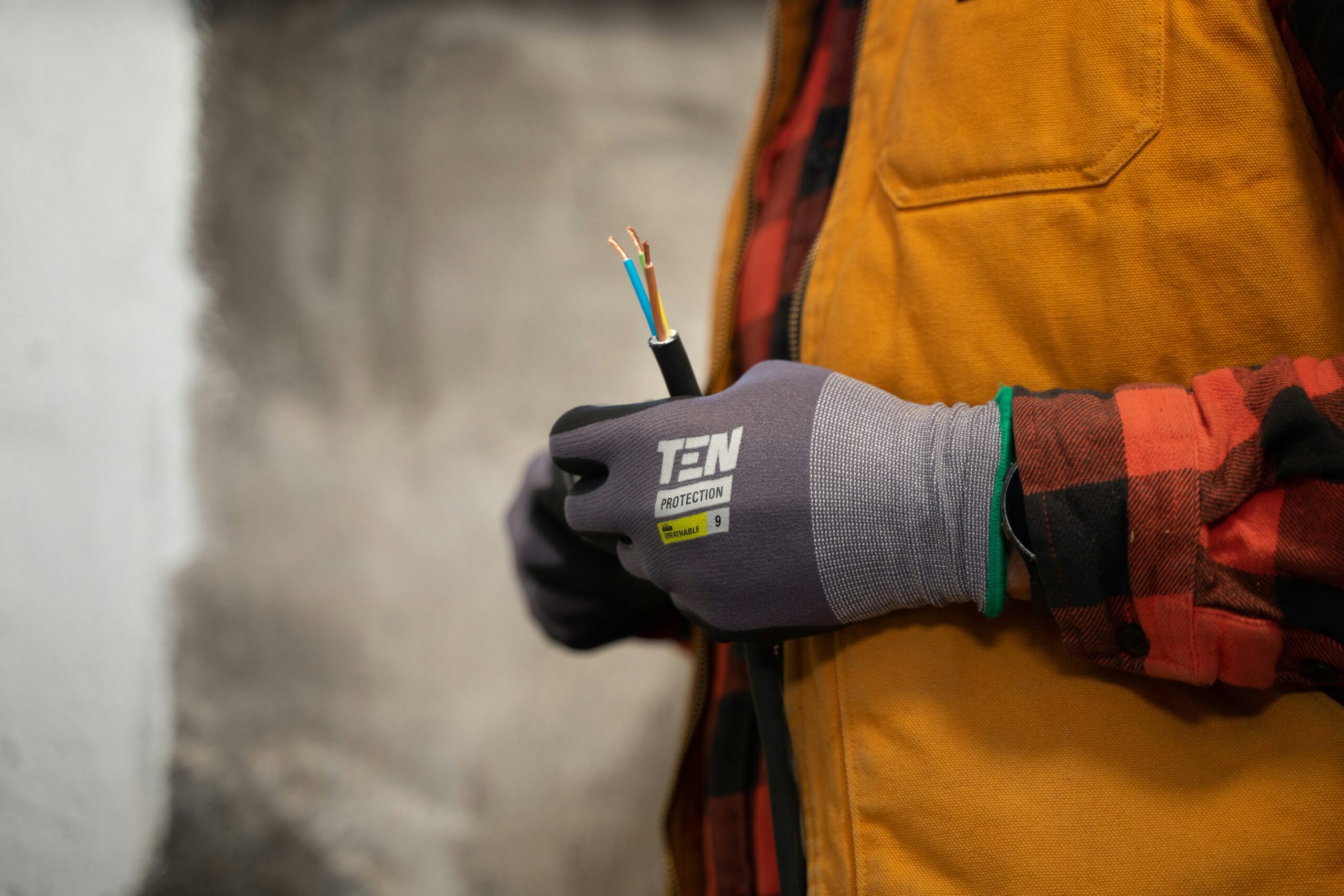 Person wearing gloves and a yellow vest holds a cut electrical wire with exposed copper and colored insulation ends in front of a blurred background.