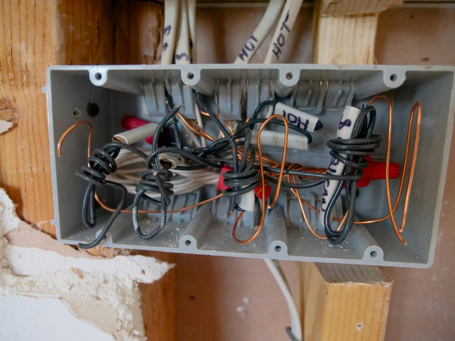 An electrical junction box with exposed wires, some twisted together and some capped, inside a wall with visible wooden studs and plaster damage.