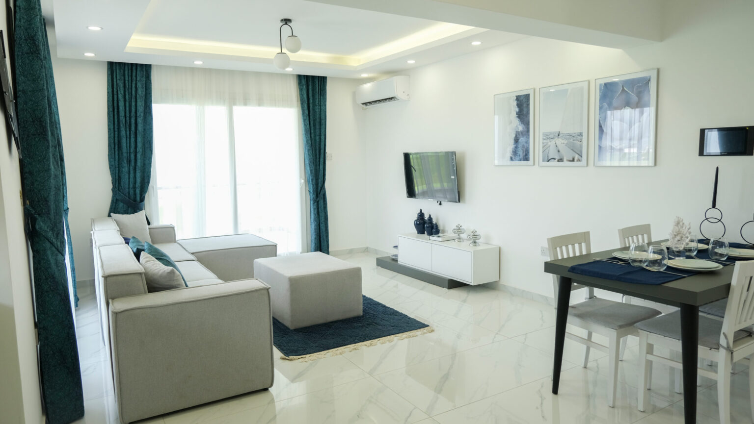 Modern living and dining area with white walls, grey furniture, blue accents, wall art, and large window with teal curtains.
