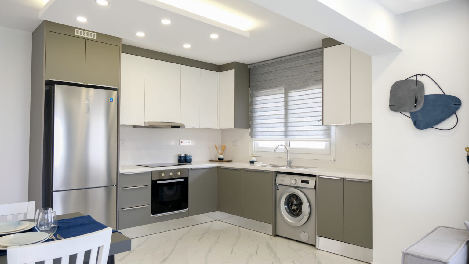 Modern kitchen with grey and white cabinets, stainless steel appliances, washing machine, induction stove, and window with grey blinds. Dining table with blue placemats in the foreground.