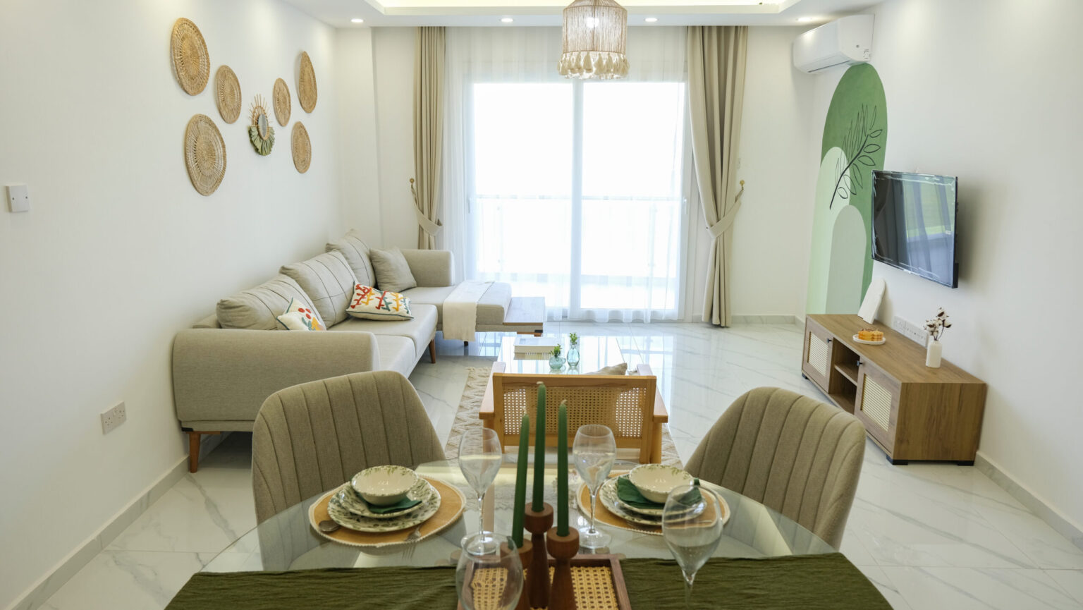 Modern living and dining room with neutral tones, a beige sofa, wall-mounted TV, dining table set for four, large window with curtains, and decorative wall accents.