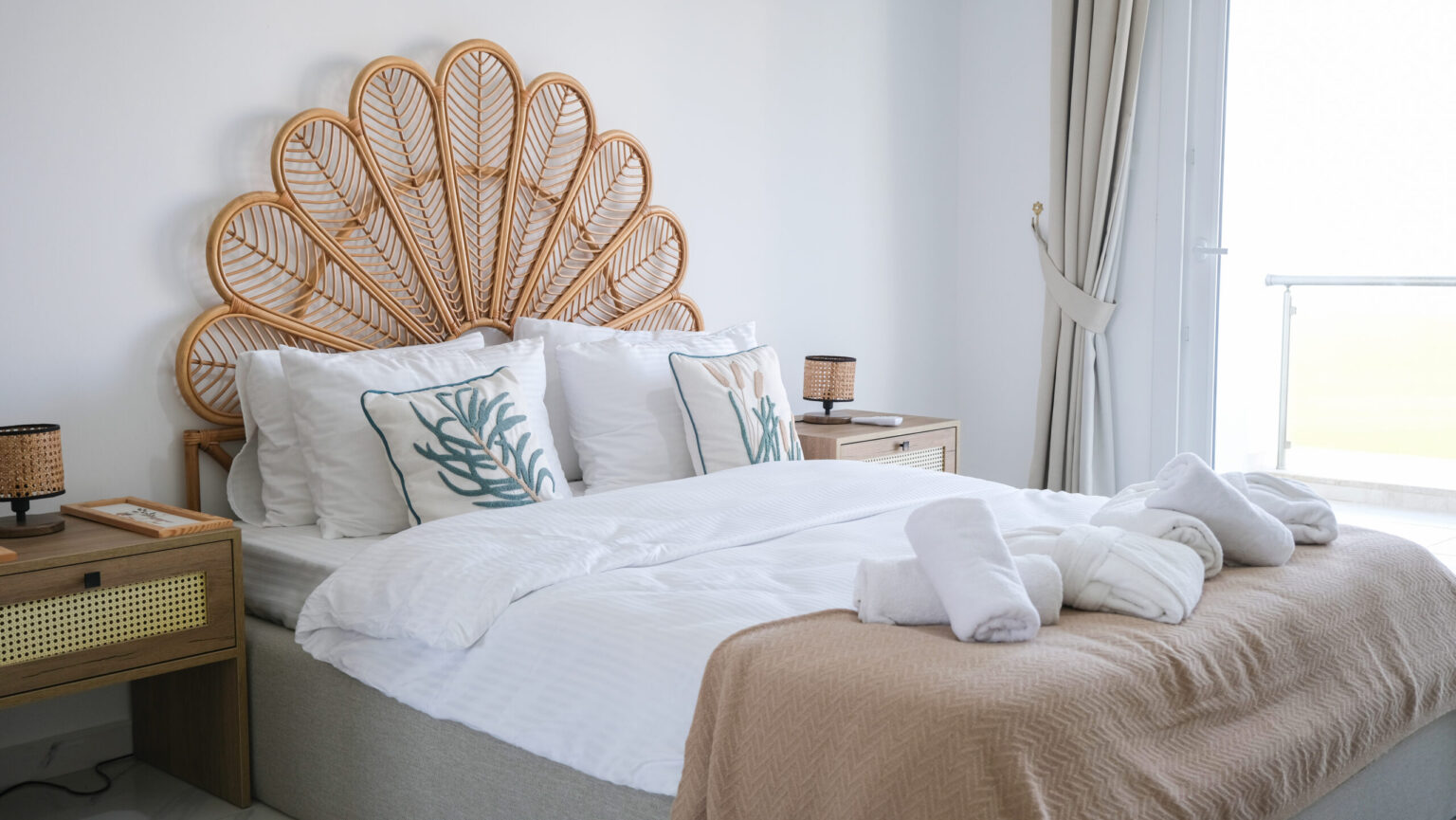 A neatly made bed with white linens, decorative pillows, and a beige throw blanket stands between two nightstands; towels are folded on the bed. The room is bright with natural light.