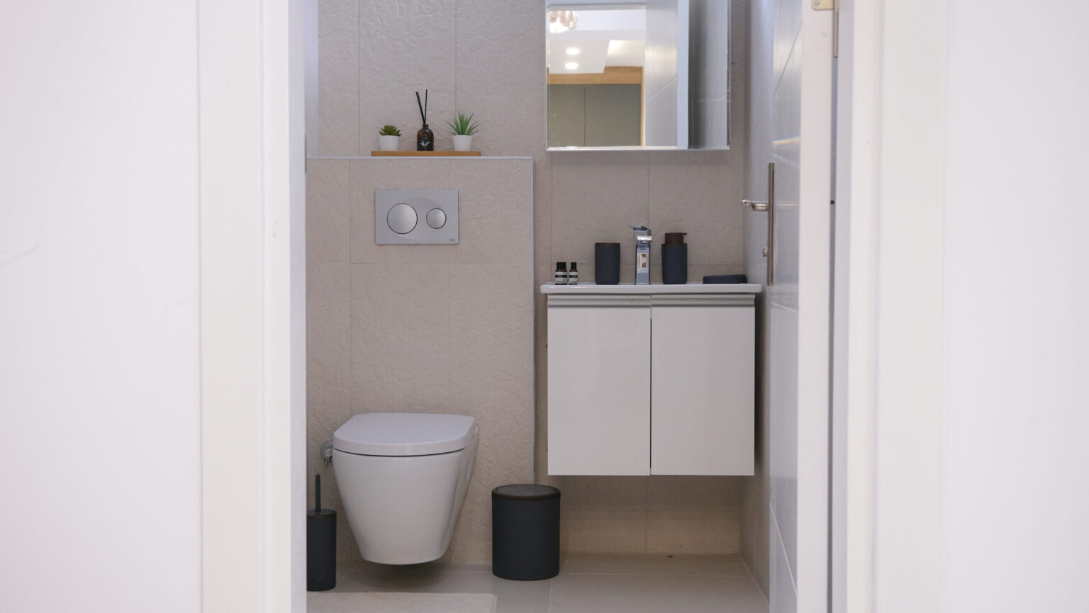 Modern bathroom with a wall-mounted toilet, white vanity, small plants, toiletries, and a mirror above the sink; neutral tones and minimalist decor.