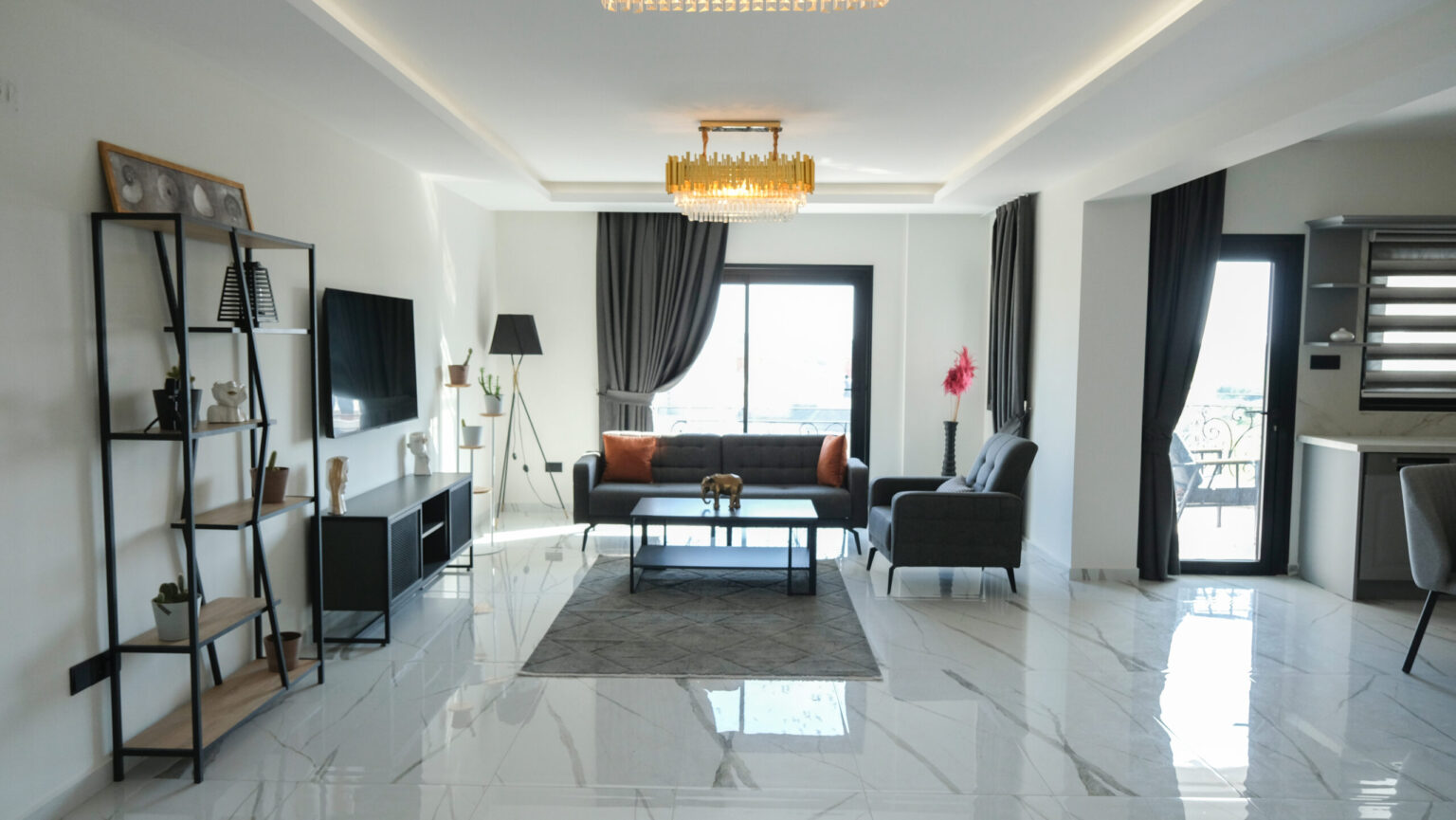 Modern living room with marble floor, gray furniture, wall-mounted TV, tall shelf, large windows with curtains, and a chandelier. Kitchen area visible on the right.