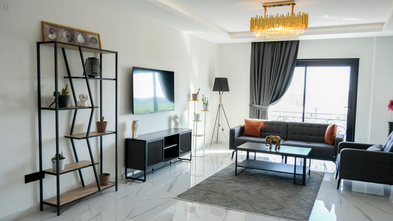 Modern living room with black and gray furniture, wall-mounted TV, metal shelving, a chandelier, and large window with dark curtains letting in natural light.