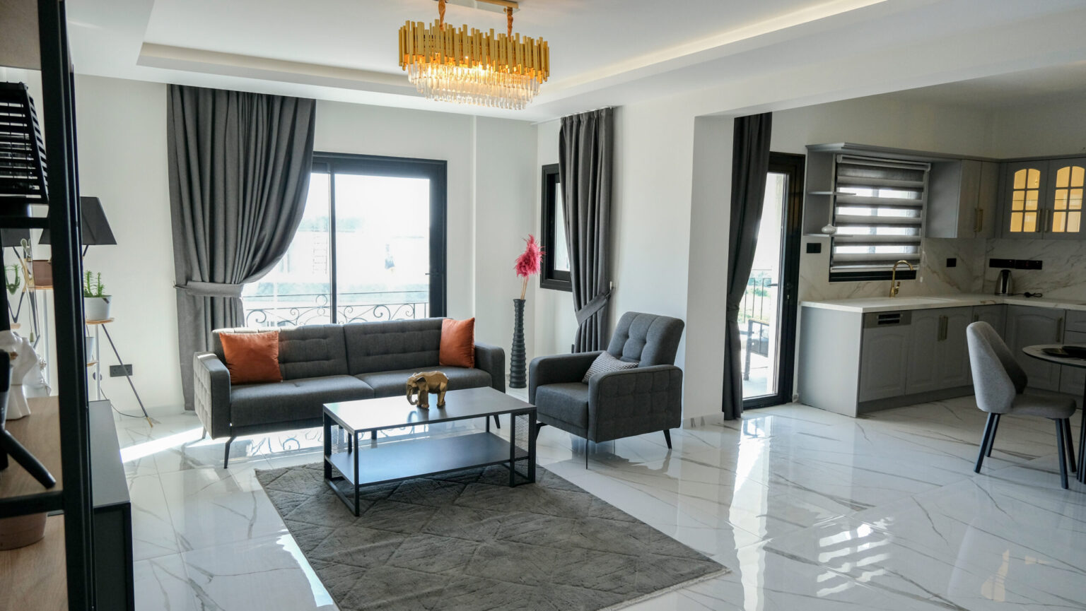 Modern living room with gray furniture, marble flooring, a chandelier, large windows with dark curtains, and an open kitchen area.