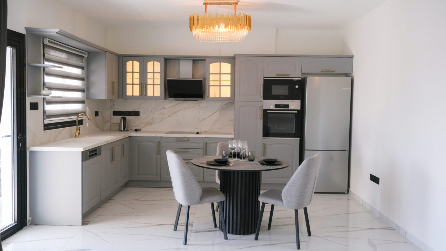 Modern kitchen with grey cabinets, marble backsplash, built-in appliances, round dining table set for four, and a chandelier overhead.