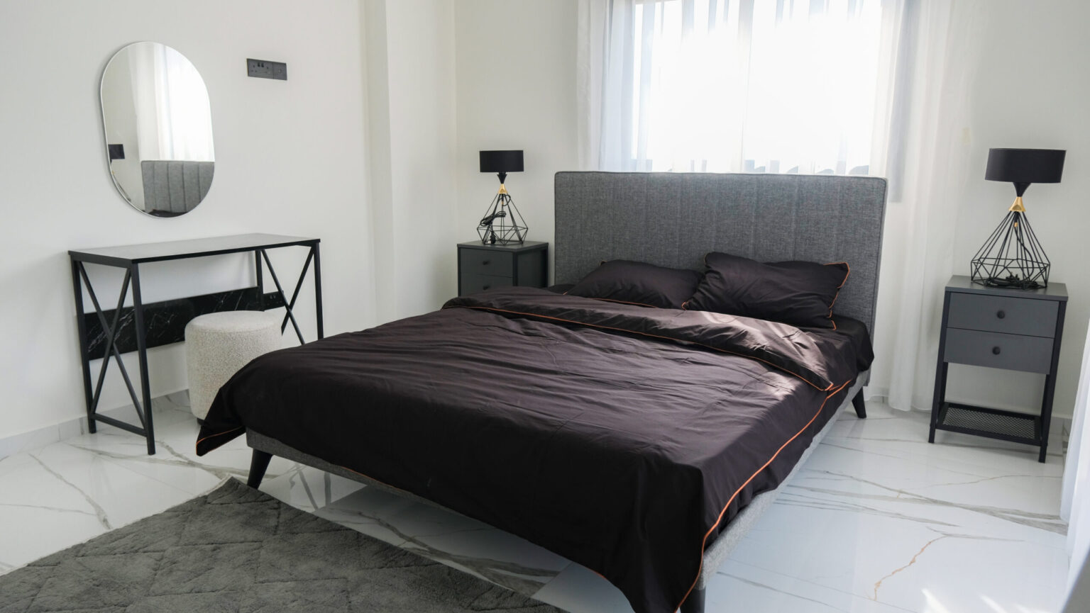 A modern bedroom with a gray upholstered bed, black bedding, two black nightstands with lamps, a vanity table with a round mirror, and a white ottoman on a marble floor.