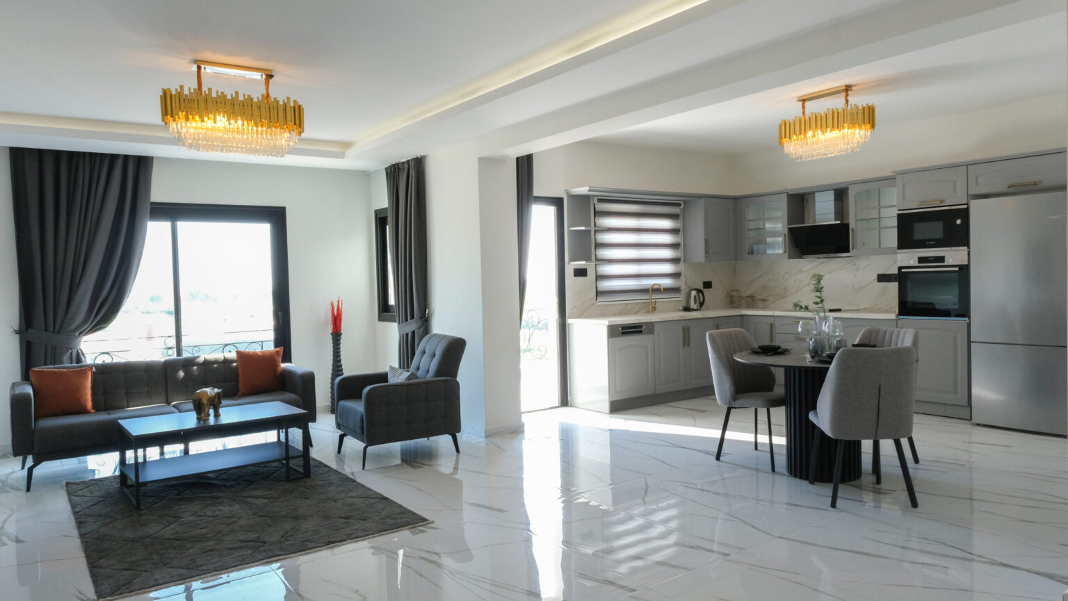 Modern open-plan living room and kitchen with grey furniture, marble floor, large windows, and two gold chandeliers.