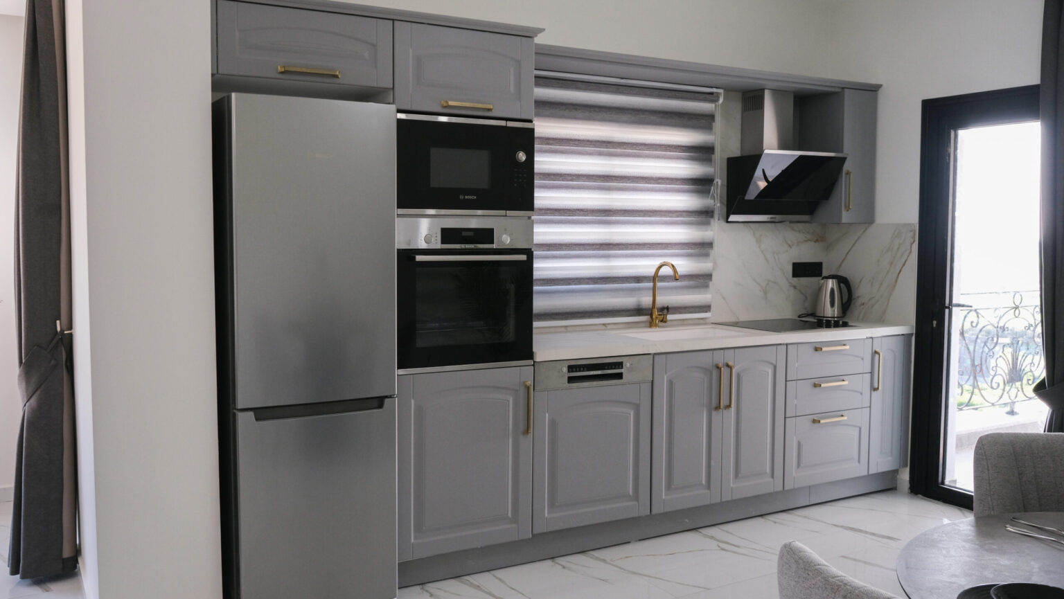 Modern kitchen with gray cabinets, built-in oven, microwave, refrigerator, electric stove, sink, kettle, and striped window blind above the countertop.