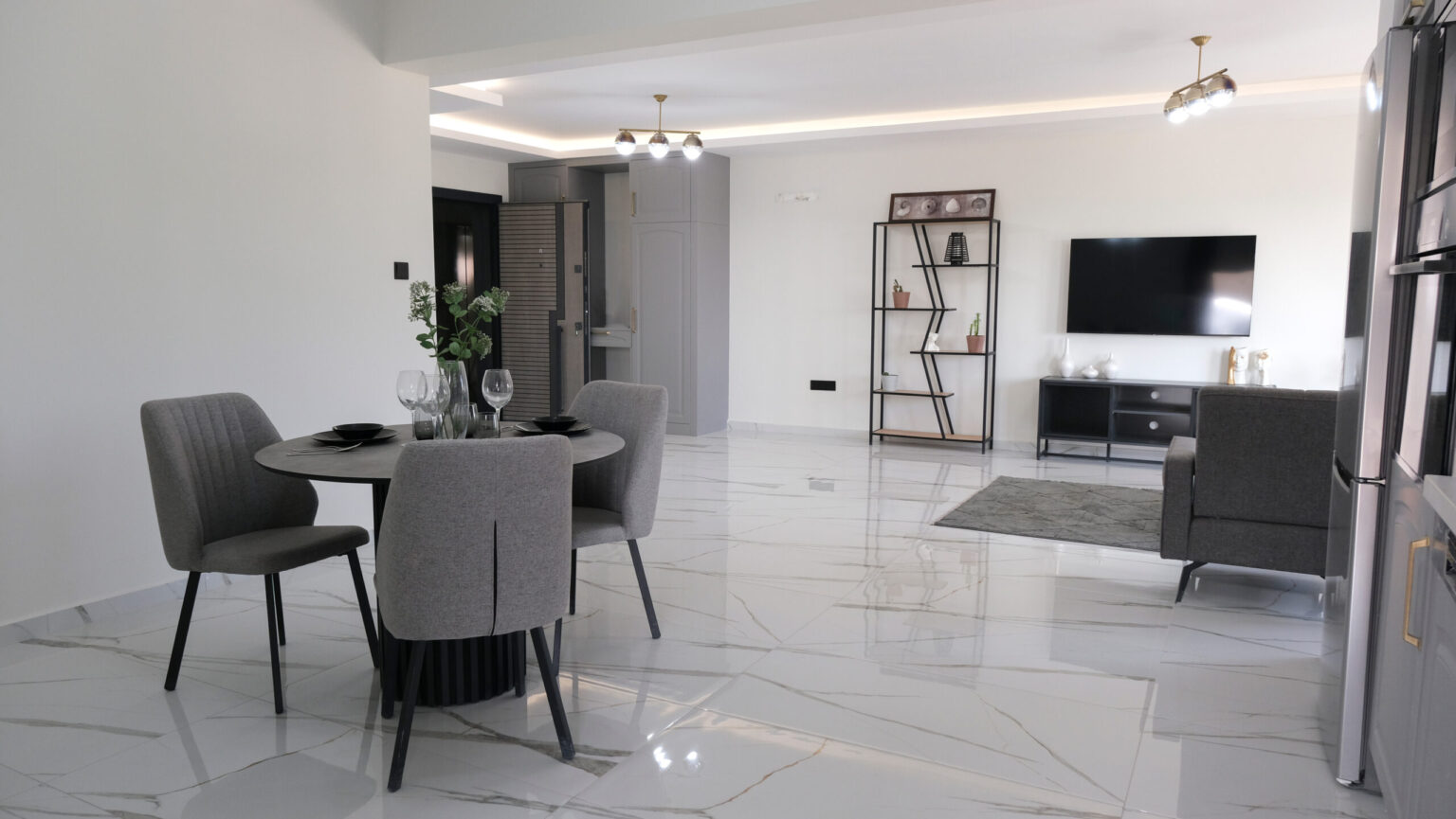Modern open-plan living space with a dining table and four chairs, a TV on a stand, a shelving unit, and marble tile flooring.