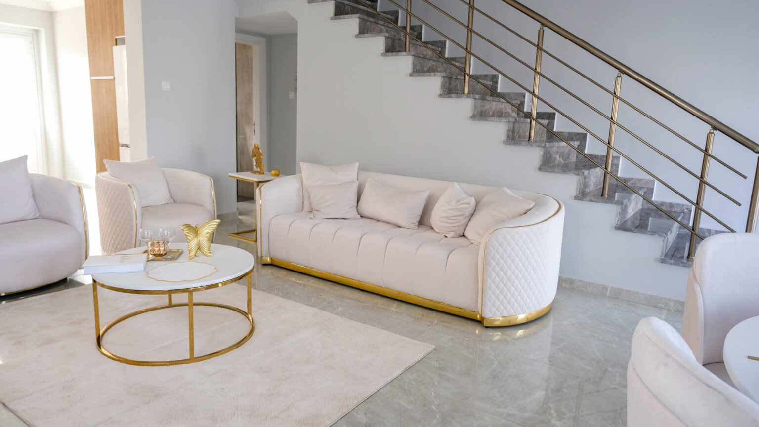 Modern living room with white sofas, a round coffee table, and a staircase with a metal railing. The decor features gold accents and light-colored flooring.