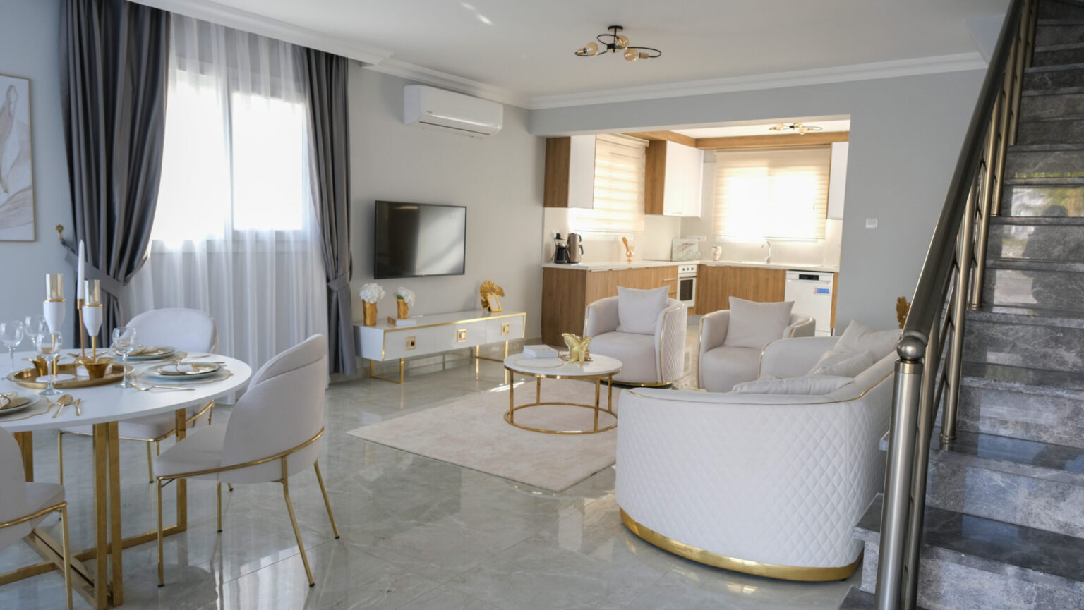 Modern open-plan living room and kitchen with white and gold furniture, marble floor, dining area, and a staircase on the right. Natural light enters through large windows.