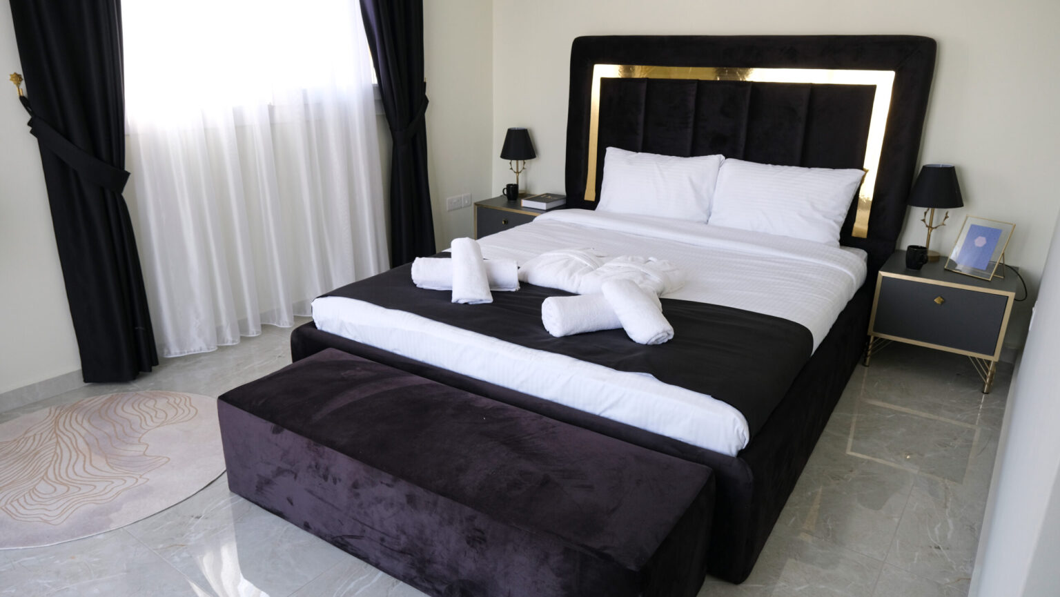 A neatly made bed with white linens, black and gold headboard, bathrobes and towels arranged on top, two nightstands with lamps, and a window with black and white curtains.