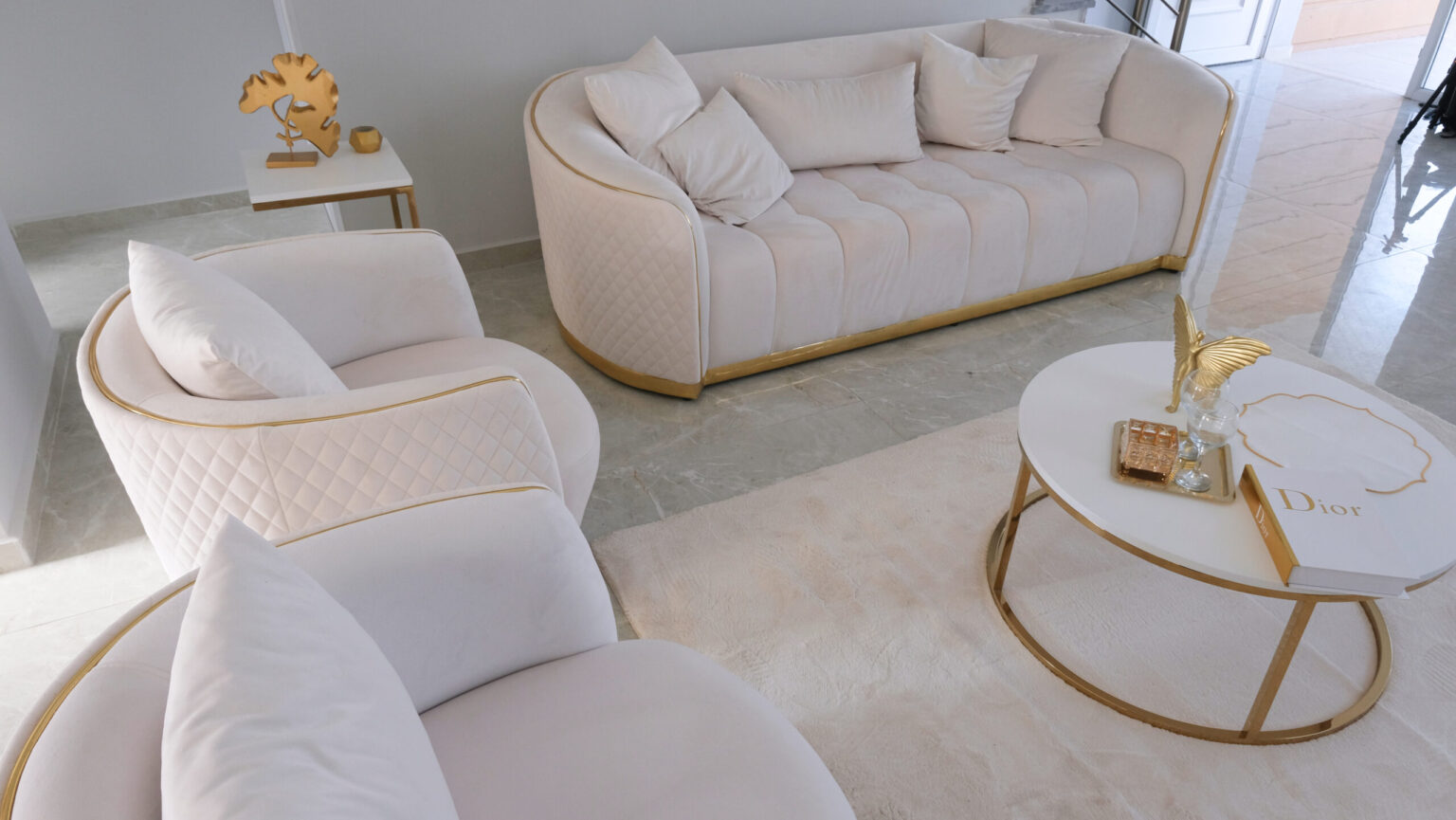 A modern living room with a white sofa, armchair, and coffee table featuring gold accents, decorative items, a book labeled "Dior," and a neutral-toned rug on a marble floor.