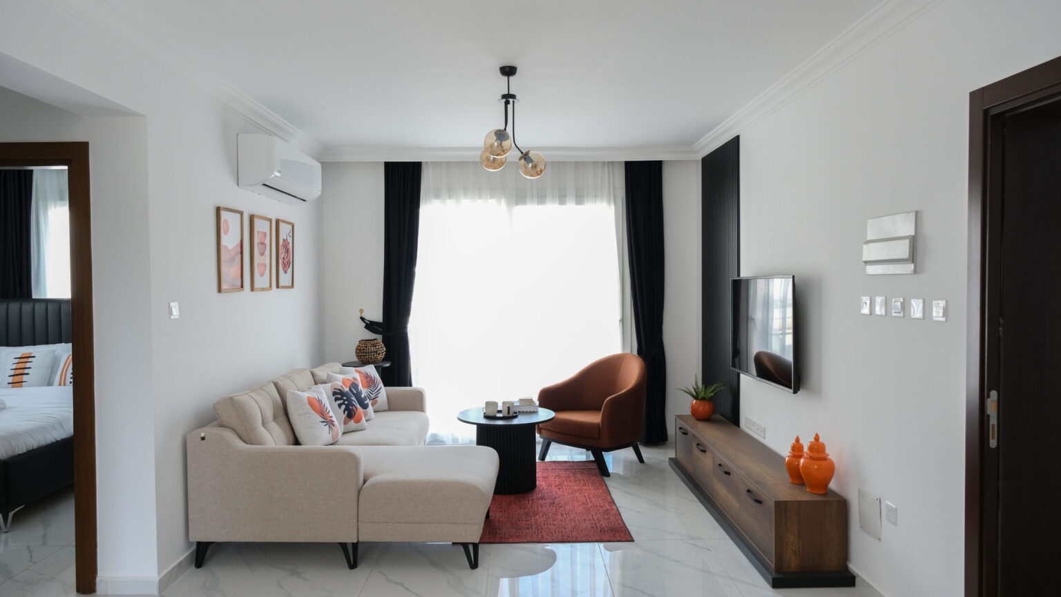 A modern living room with a beige sofa, orange chair, coffee table, TV on a wooden stand, and wall art. Large window with curtains and light fixture overhead. Bedroom visible on the left.