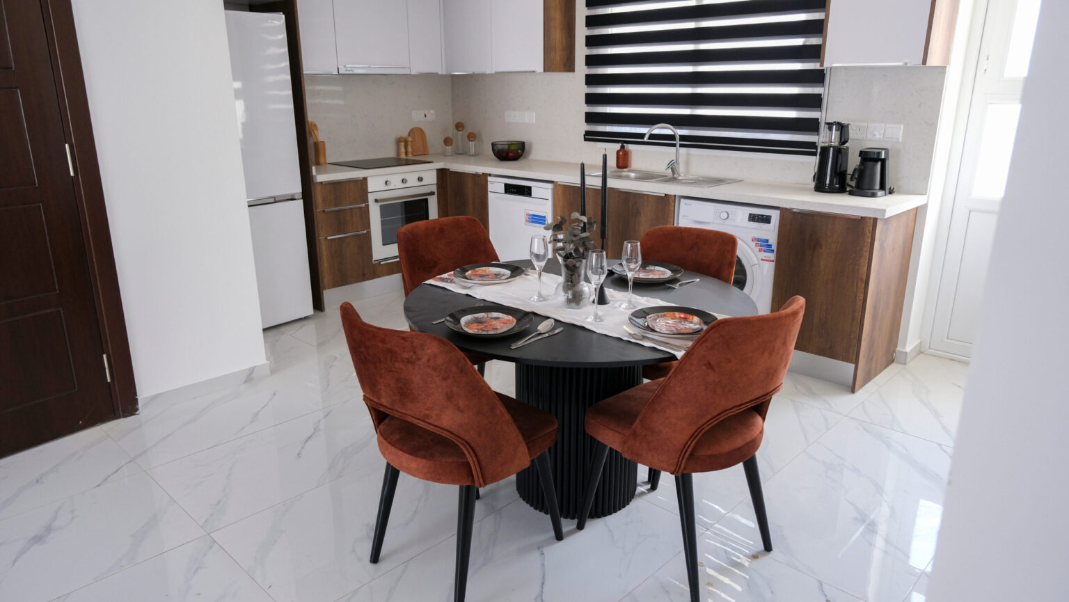 A modern kitchen and dining area with white cabinets, wood accents, four rust-colored chairs, a round black table set for four, and appliances including a fridge, oven, and dishwasher.