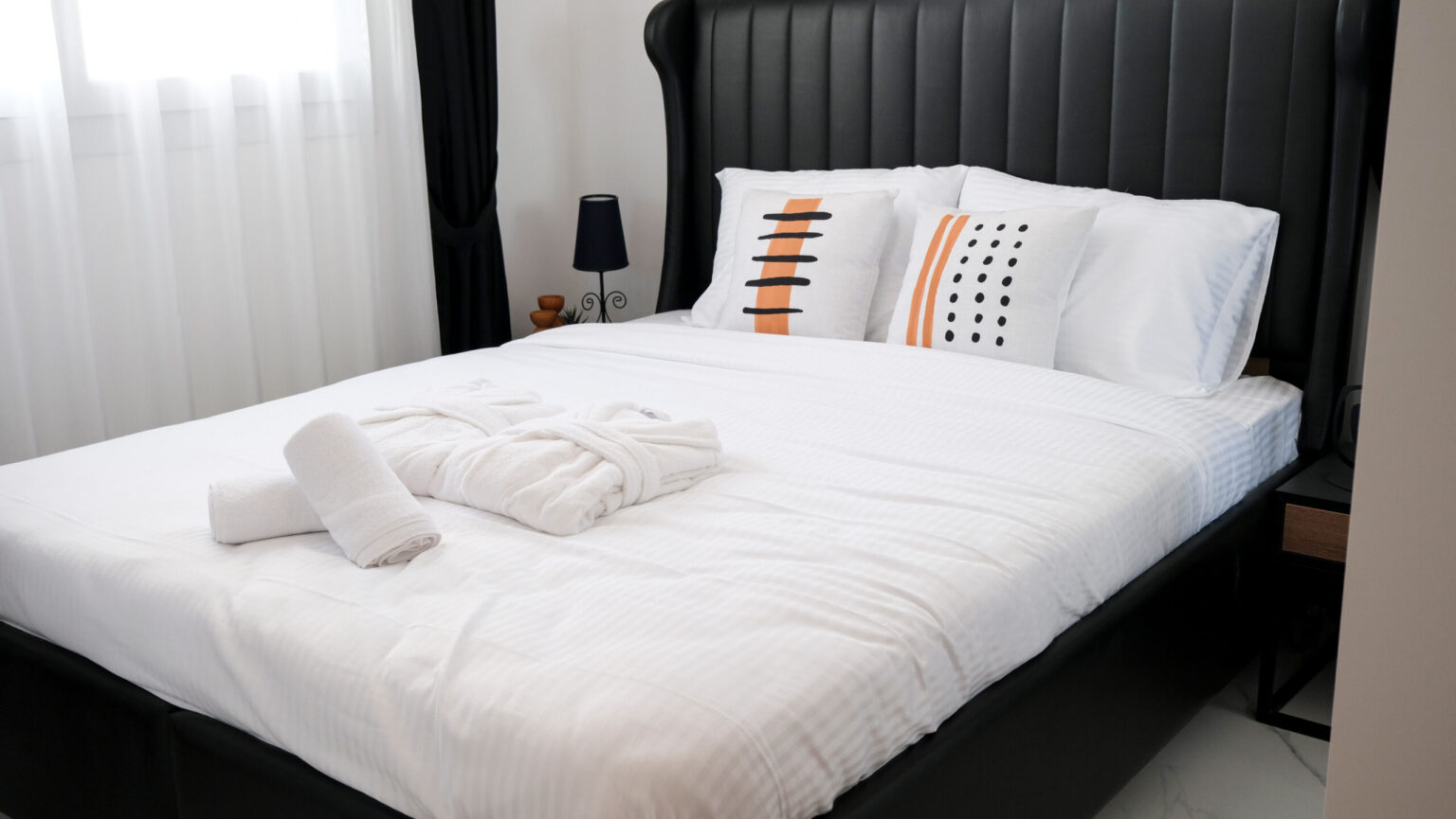 A neatly made bed with white linens, two patterned pillows, a folded bathrobe, and two towels; a bedside table with a lamp is visible.